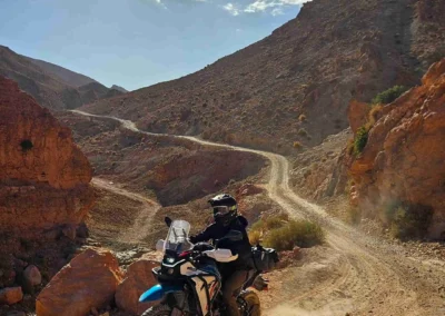 ADV rider on a dirt bike climbing a dusty switchback track in the Moroccan mountains.