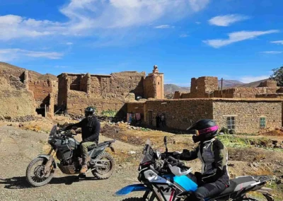 Two ADV riders on trail motorcycles stopping near old stone buildings in a remote Moroccan village.