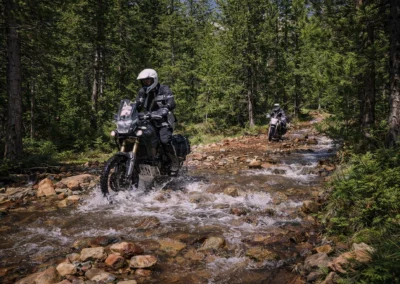 Two ADV riders crossing a rocky mountain stream in the Balkan forest.